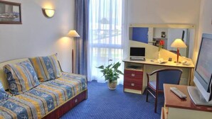 A hotel room with a sofa, a desk with a computer, a chair, a TV, and a window with curtains. - Appart'City Classic Rennes Beauregard (Rennes)