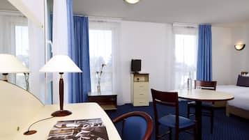 Ein Hotelzimmer mit einem Bett, einem Schreibtisch mit Lampe, einem Sessel, einem kleinen Tisch, einem Fernseher und einer Kommode.