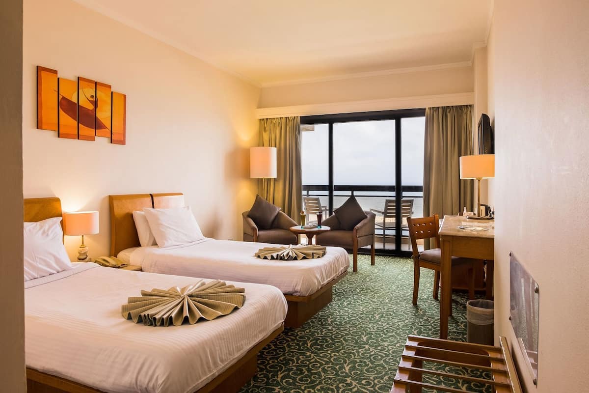 A hotel room with two beds, a desk, chairs, a view of the sea, and artwork on the wall.