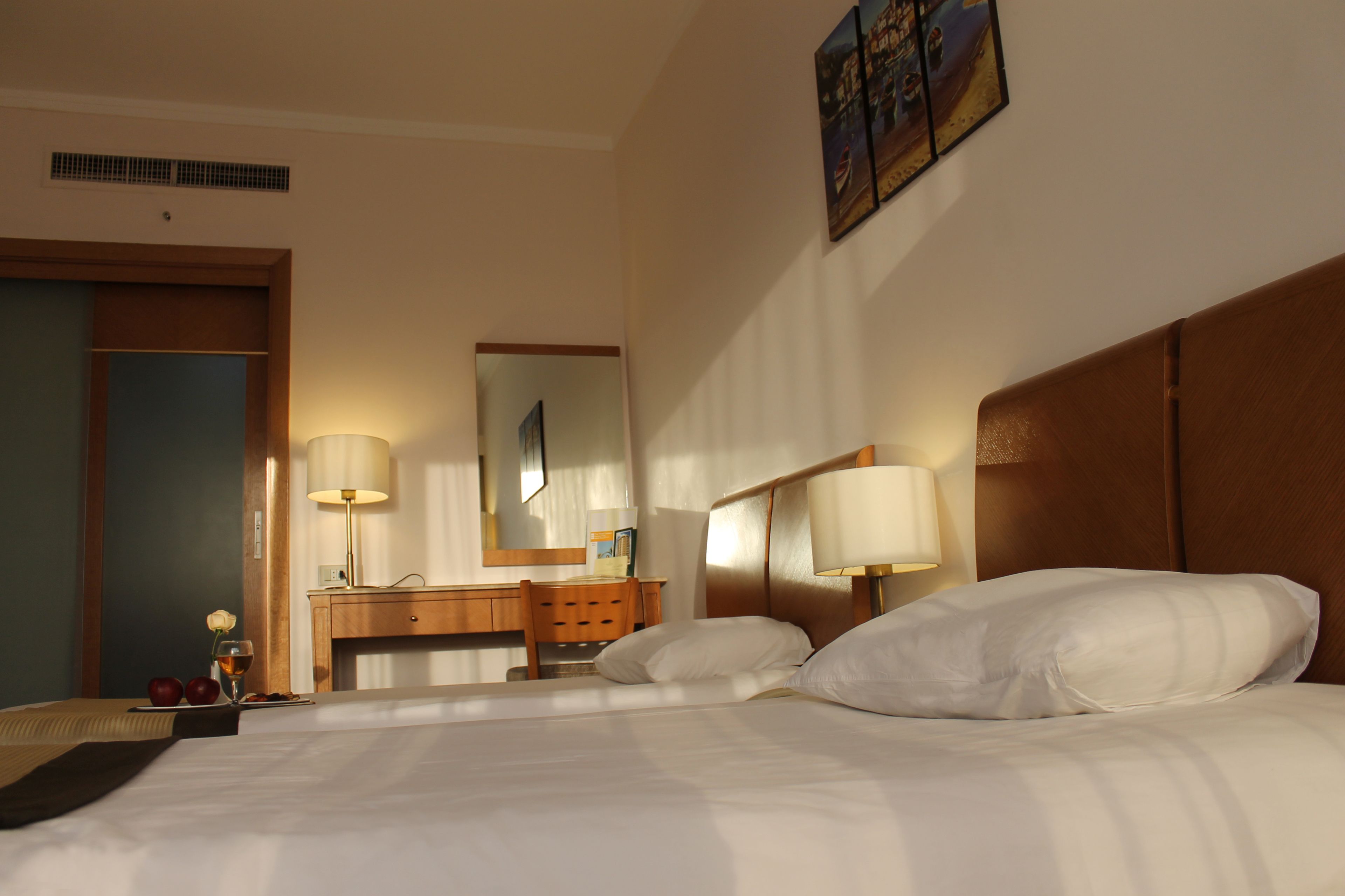 A hotel room with a bed, two bedside lamps, a wooden headboard, a desk with a chair, a mirror, and a wall-mounted artwork.