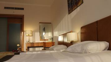 A hotel room with a bed, two bedside lamps, a wooden headboard, a desk with a chair, a mirror, and a wall-mounted artwork.