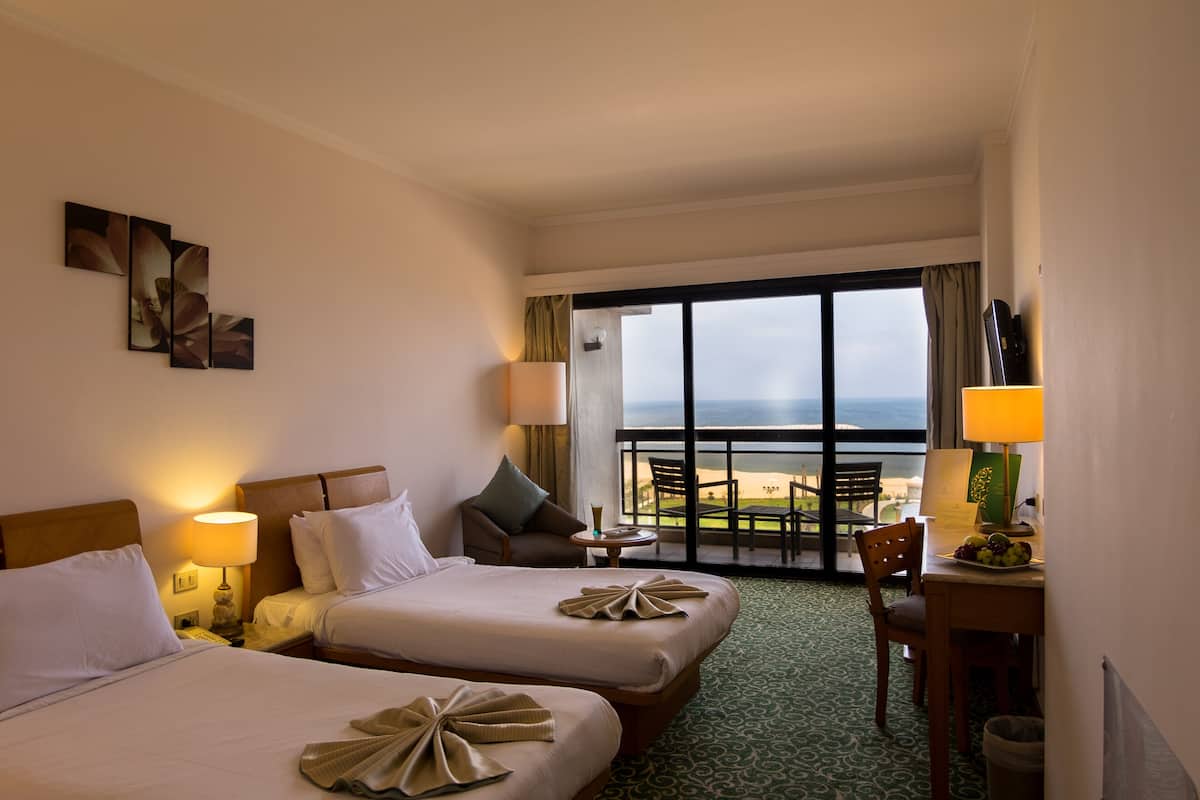 A hotel room with two beds, a balcony with a view, and a small table with a lamp.