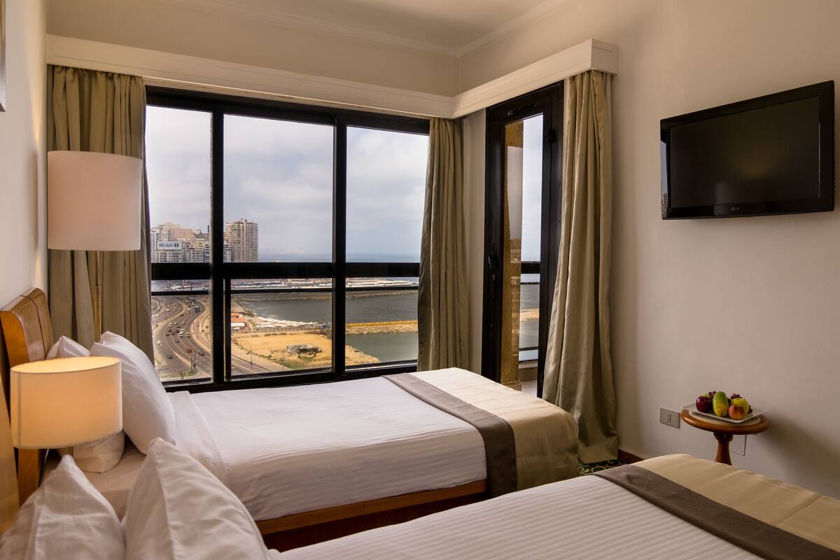 A hotel room with two beds, a TV on the wall, and a view of a bridge and water.