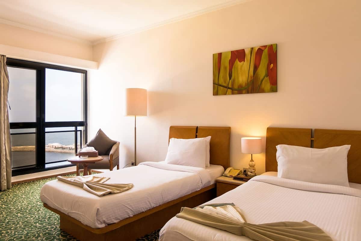 A hotel room with two beds, a chair, a table, a lamp, a painting, and a view of the sea.