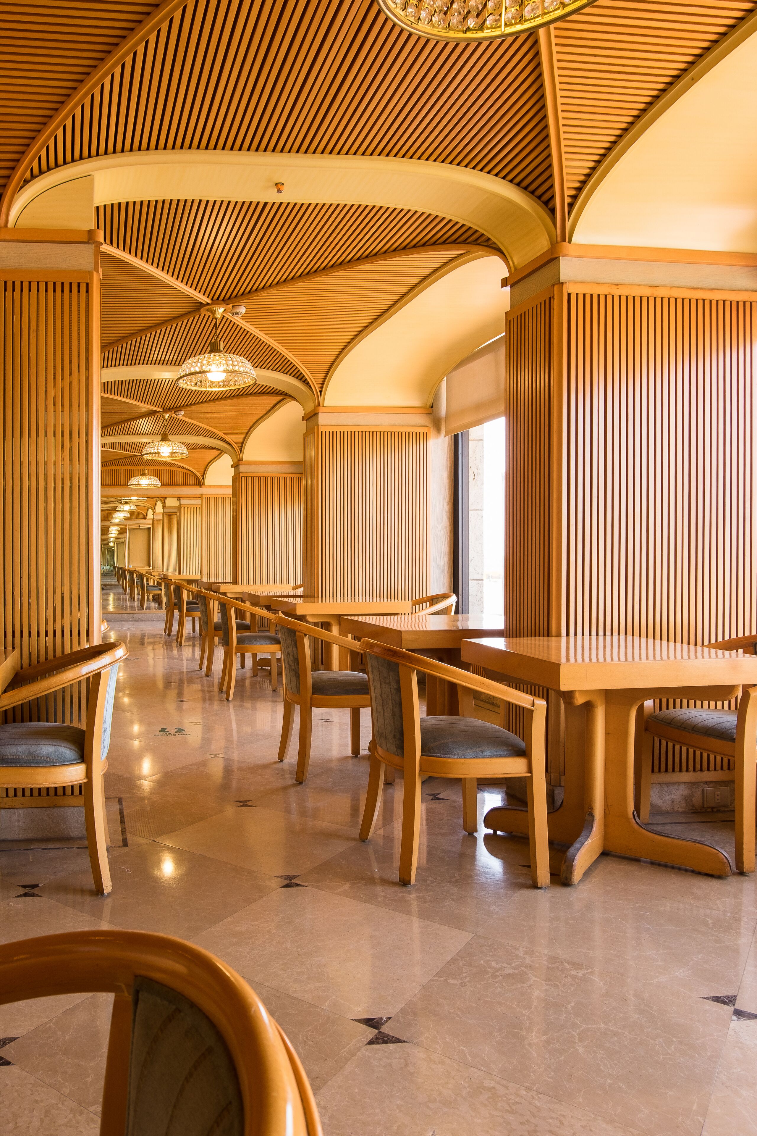 Un comedor amplio y moderno con mesas y sillas de madera, que presenta un techo alto con un diseño original de listones y una araña.