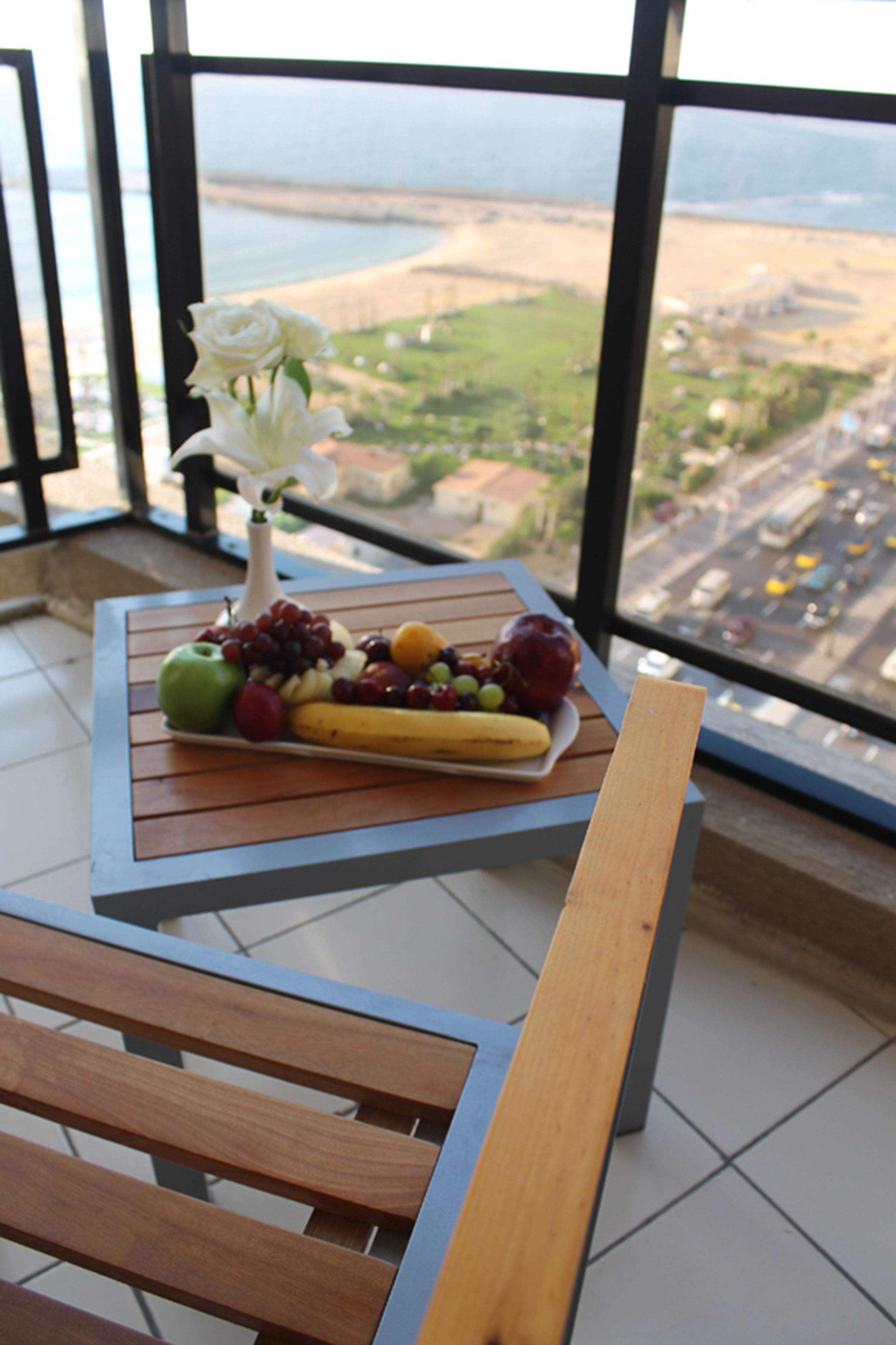 A table with a fruit platter and a white vase with a single white flower, overlooking a coastal view.