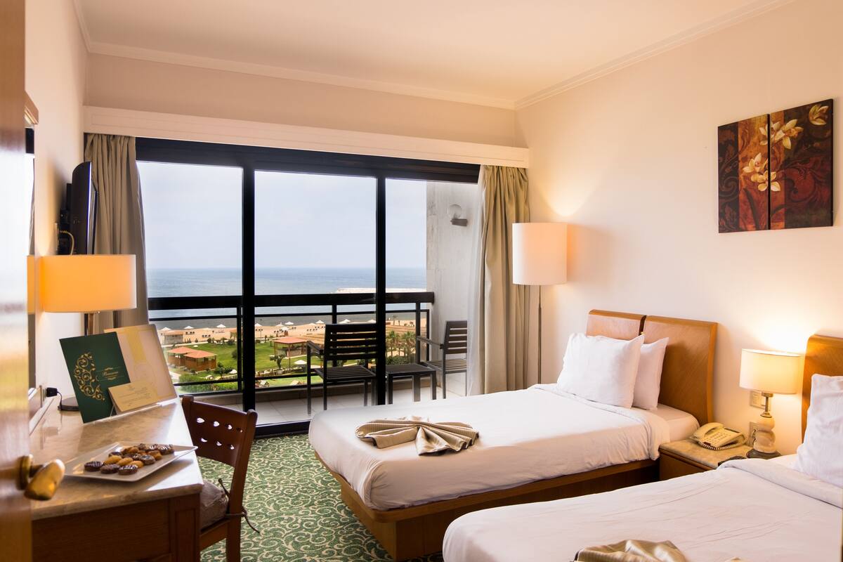 A hotel room with two beds, a balcony with a view, a desk with a lamp, and a painting on the wall.