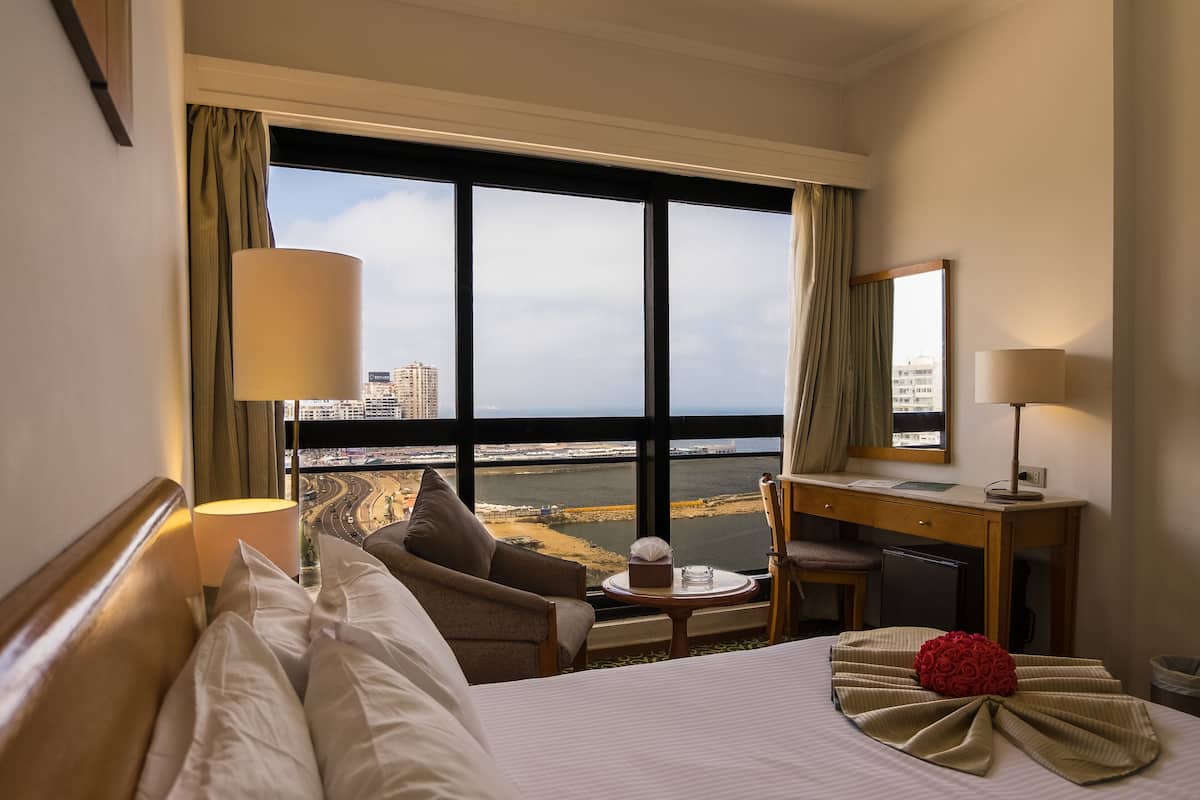 A hotel room with a large window, a bed, a desk, and a chair.