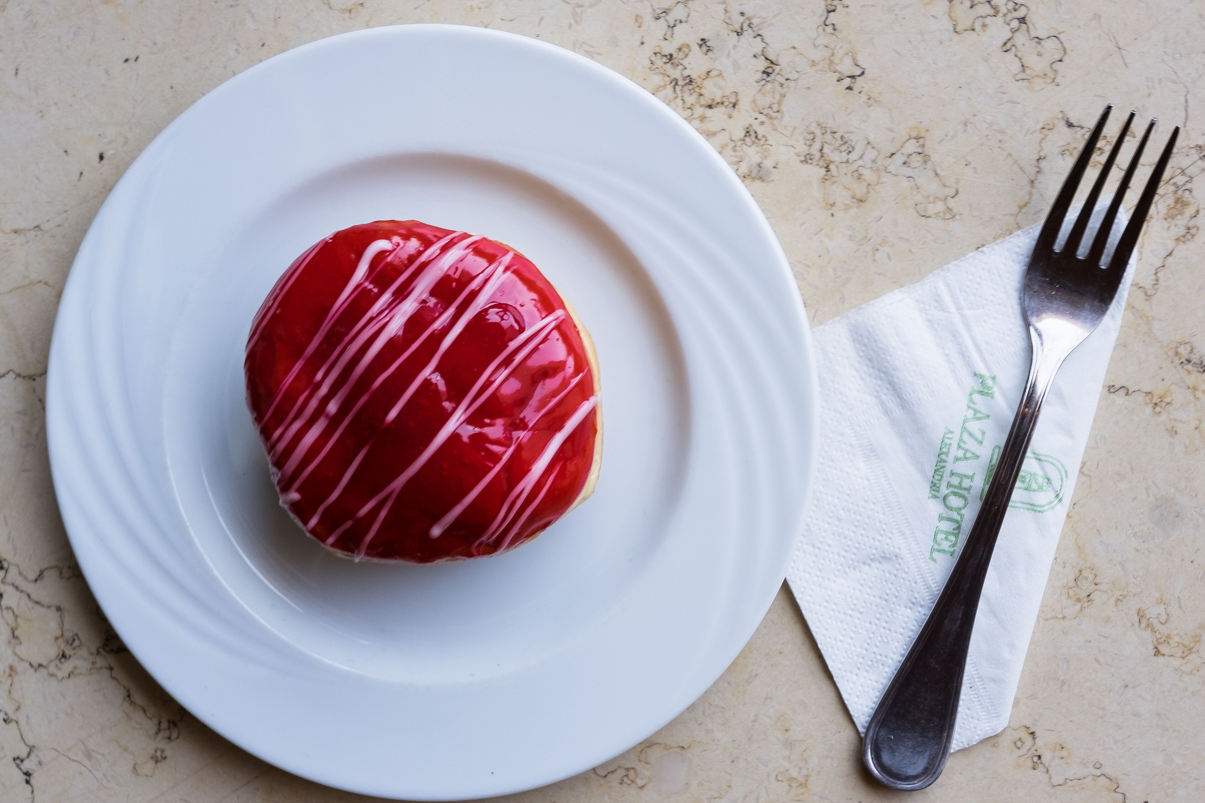 A single red donut with white icing on a white plate, accompanied by a fork and a napkin with a logo.