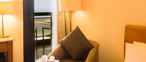 A hotel room with a bed, a chair, a small table with a tissue box, and a view of the sea through an open door.
