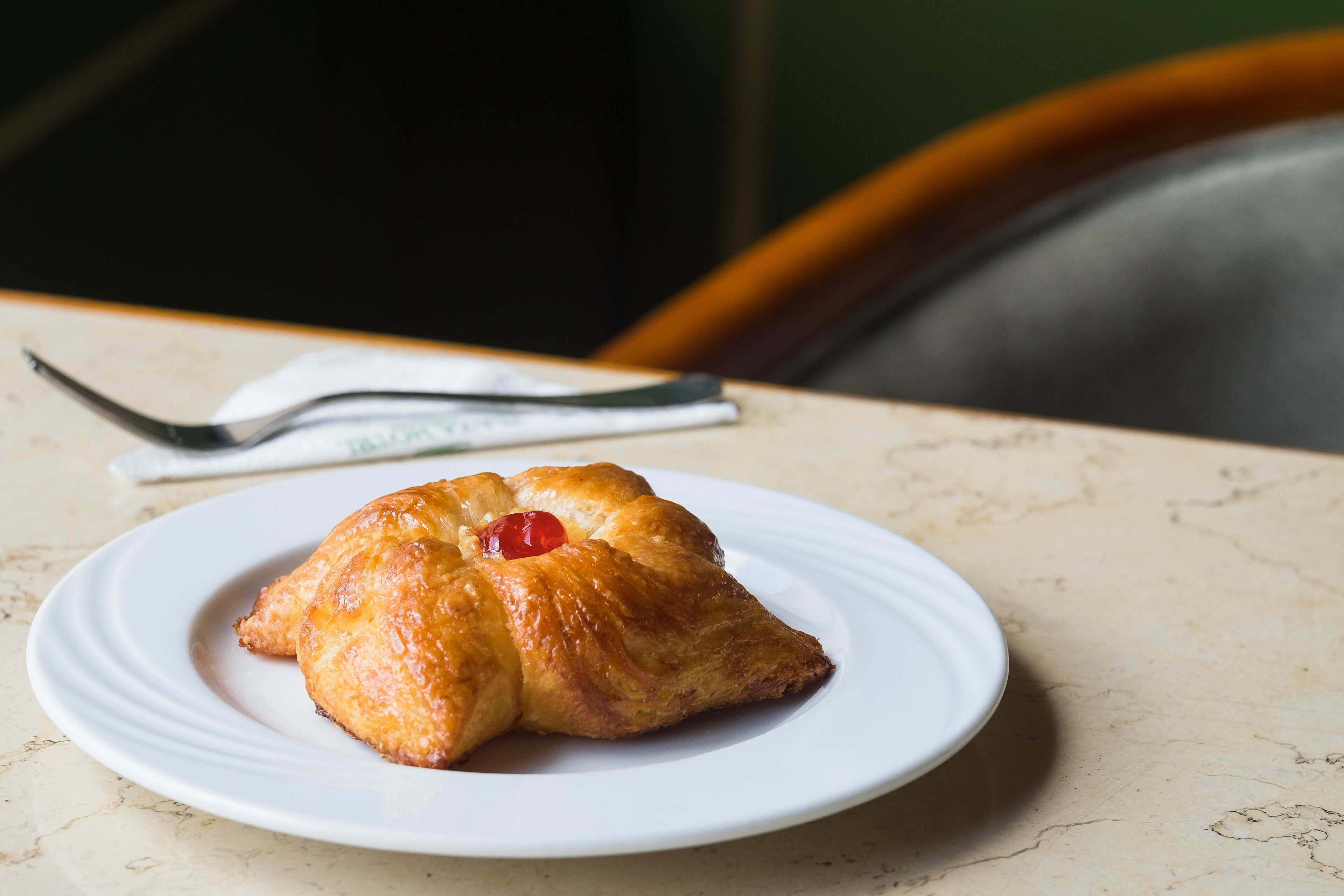 A croissant on a white plate with a cherry on top, placed on a marble table.