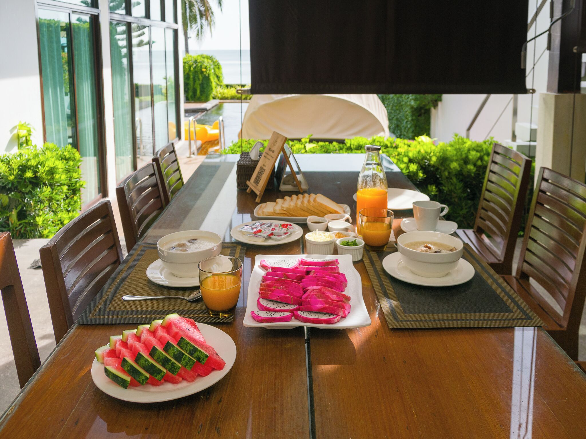 A breakfast table set with various dishes, including fruit, bread, and beverages, in a sunny outdoor dining area.