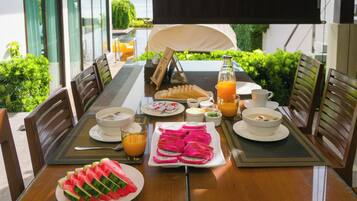 A breakfast table set with various dishes, including fruit, bread, and beverages, in a sunny outdoor dining area.