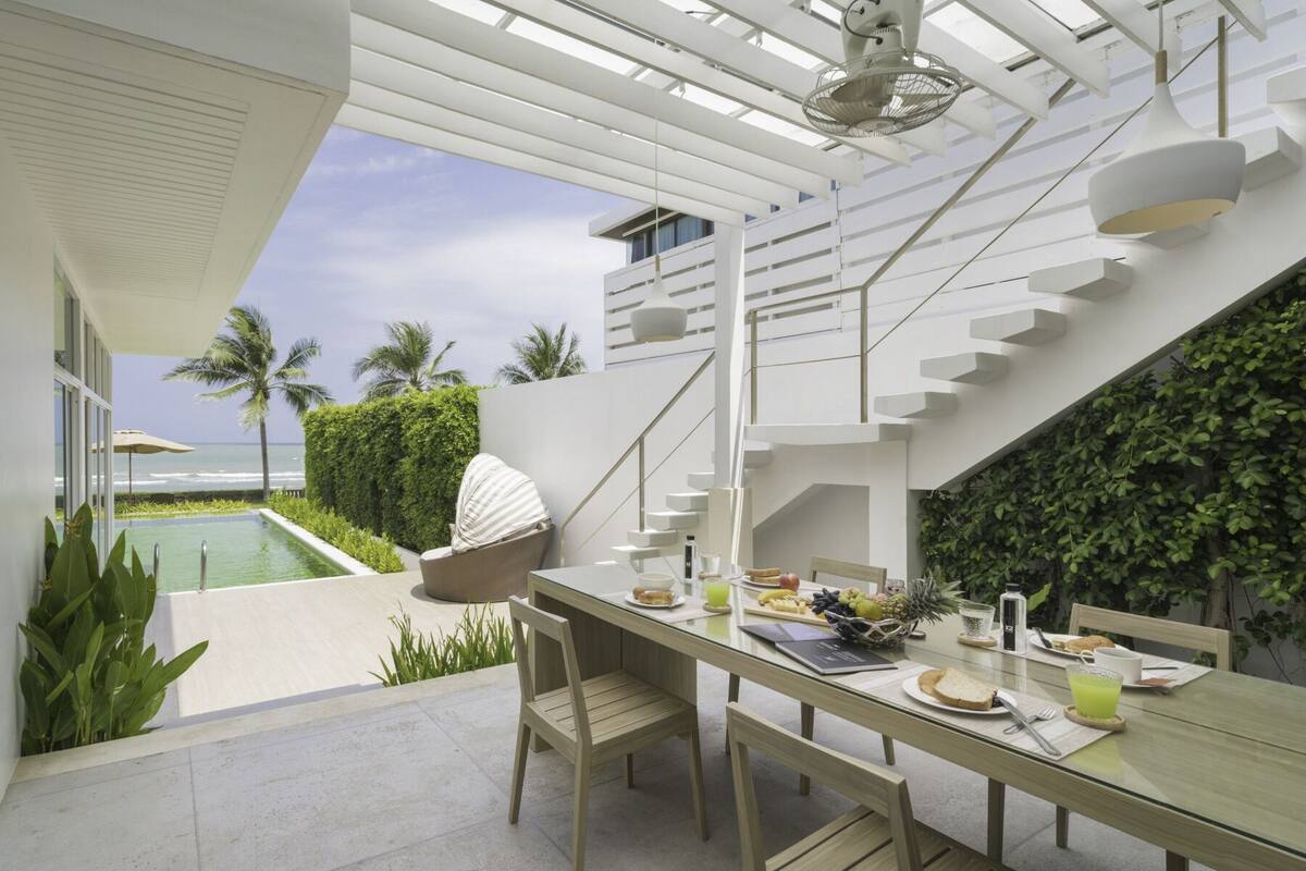 A modern outdoor dining area with a glass table, chairs, and a view of the ocean.