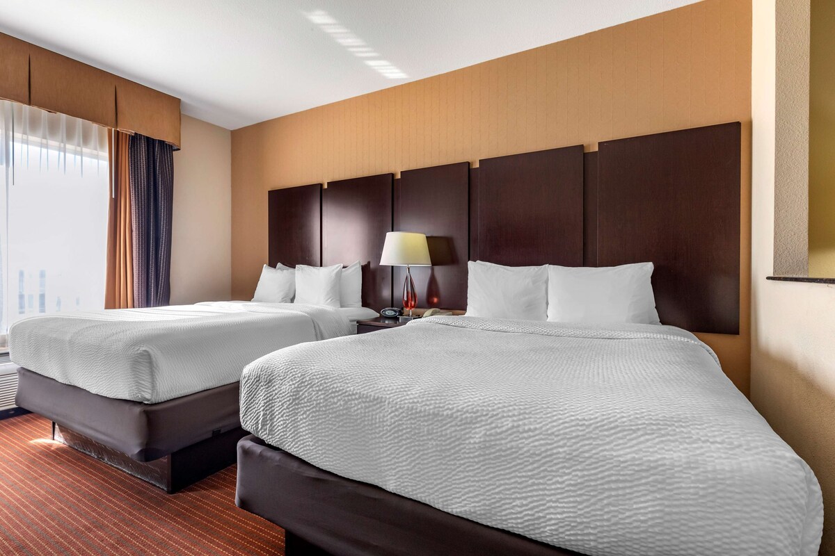 A hotel room with two beds, a large window with curtains, a nightstand with a lamp, and a wooden headboard.