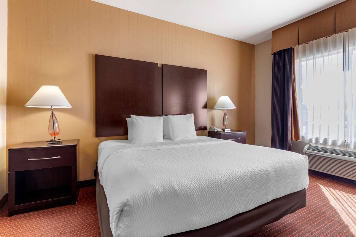 Una habitación de hotel con una cama grande, dos mesitas de noche con lámparas, un teléfono en la pared y una ventana con cortinas.