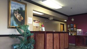 A reception area with a wooden counter, a framed painting of birds, a large potted plant, and a wall-mounted information board. - Holiday Lodge (Pittsburg)