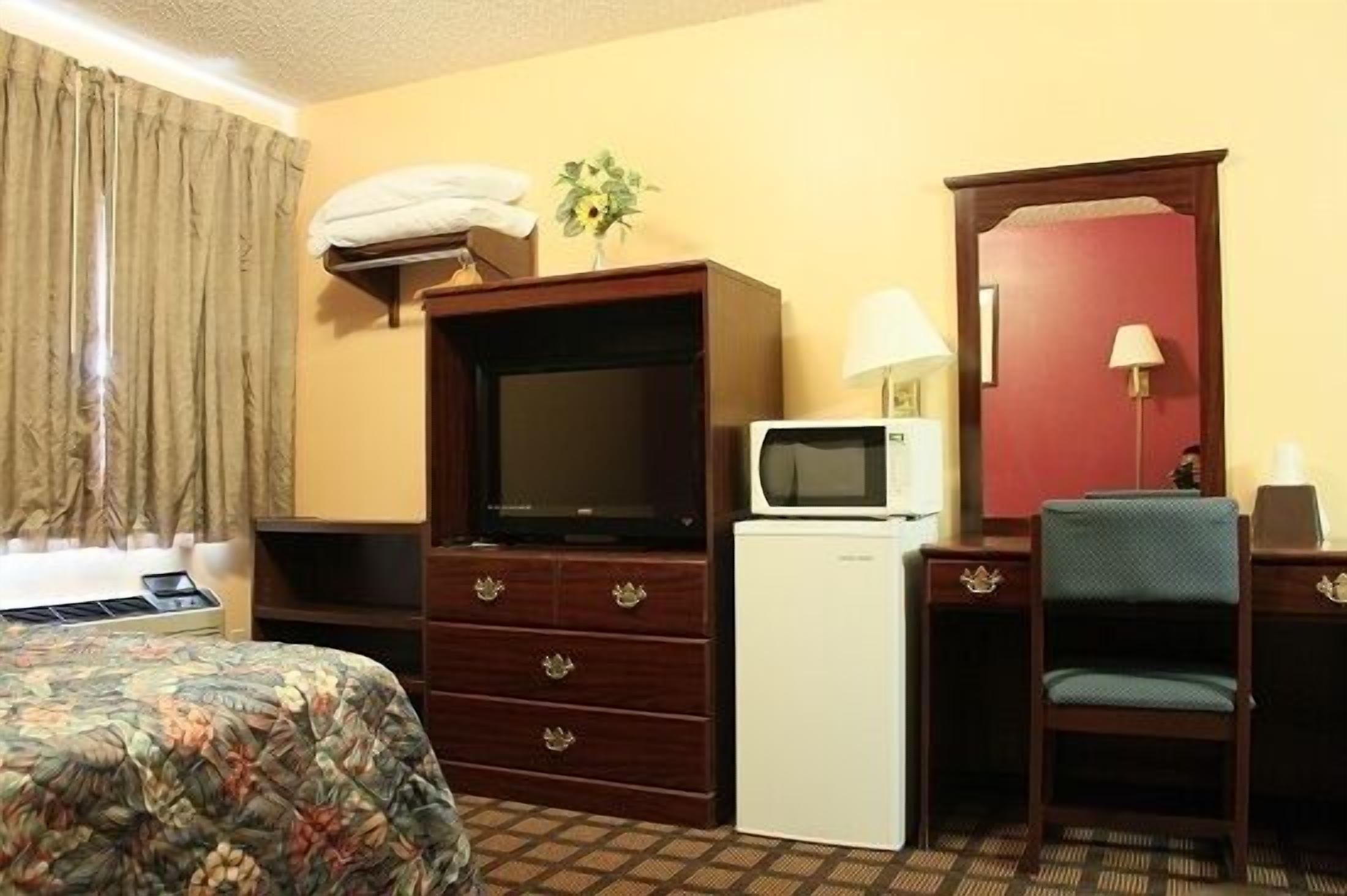 A hotel room with a bed, a desk with a chair, a microwave, a television, a small refrigerator, and a lamp.