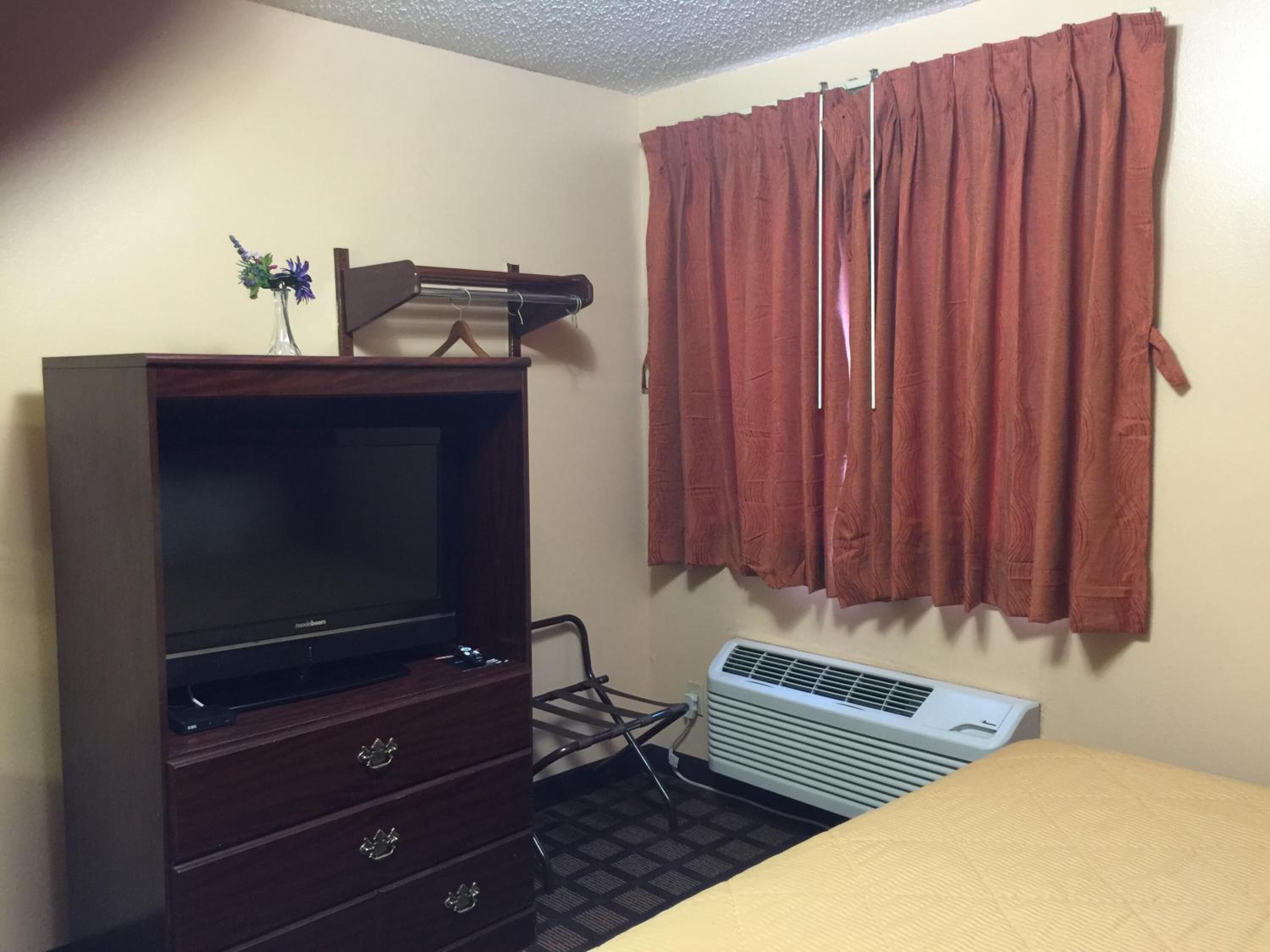 A room with a wooden dresser, a television, a bed, and a window with red curtains.