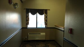 A narrow hallway with patterned carpet, a window with decorative curtains, and a wall-mounted air vent. - Holiday Lodge (Pittsburg)
