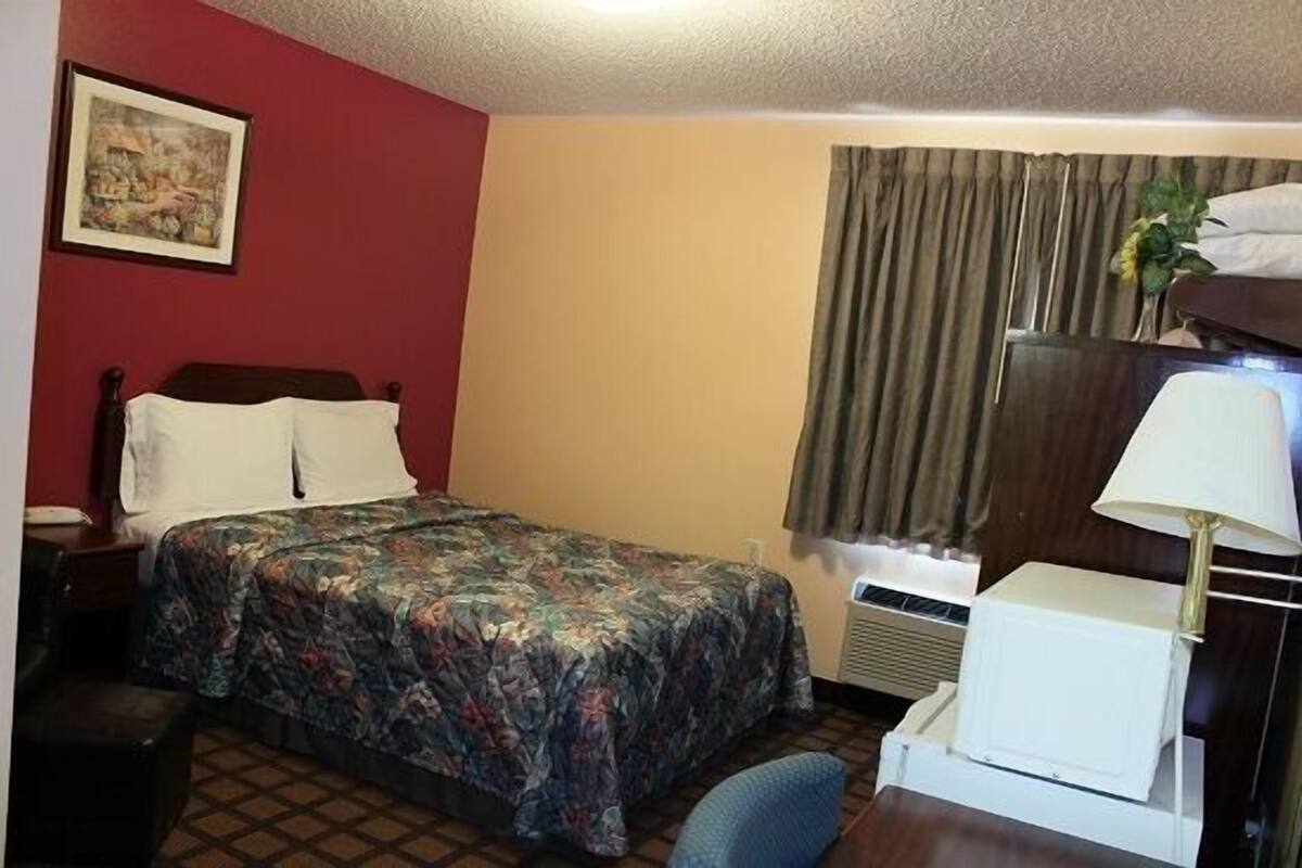Ein Hotelzimmer mit Bett, Schreibtisch, Stuhl, Lampe, einem Bild an der Wand und einem Fenster mit Vorhängen.
