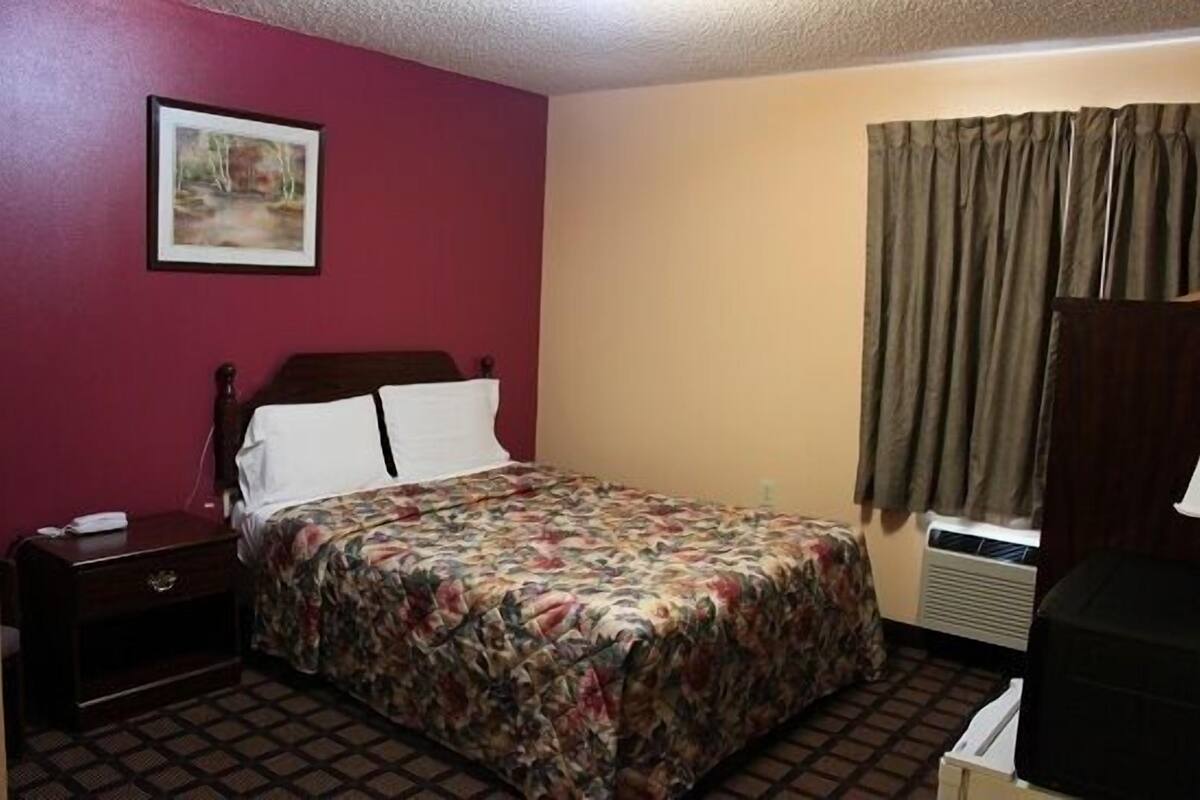 Ein Hotelzimmer mit Bett, Nachttisch, Kommode, einem Bild an der Wand und einem Fenster mit Vorhängen.