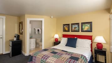 A hotel room with a bed, two bedside tables with lamps, a small desk with a lamp, a mirror, and a door leading to a bathroom.