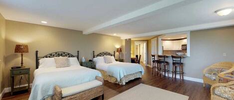 A bedroom with two beds, a small table, and a chair. There is a kitchenette in the background.