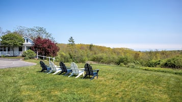 A grassy area with four Adirondack chairs facing a white building with a porch.