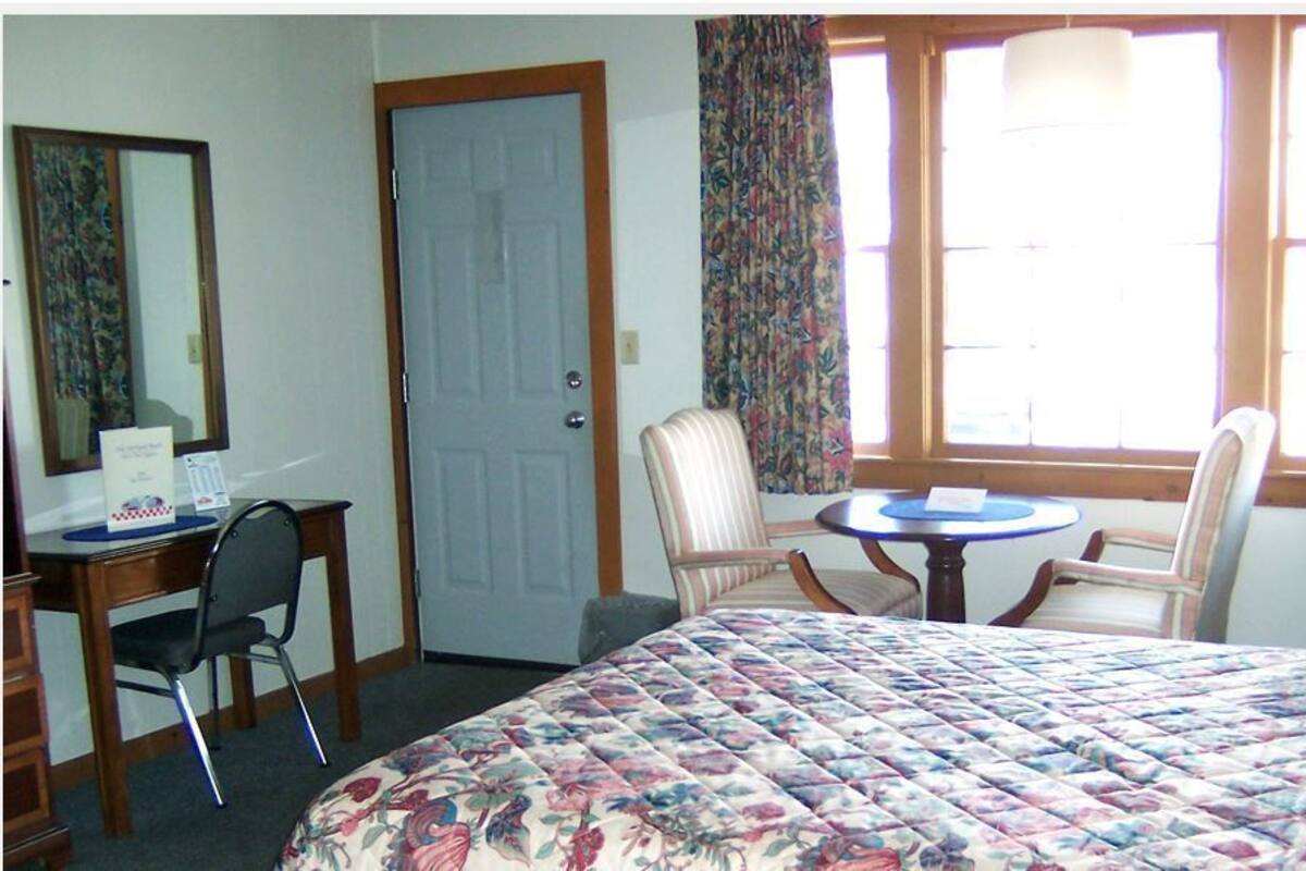 A hotel room with a bed, a desk with a chair, a small table with two chairs, a door, and a window with curtains.