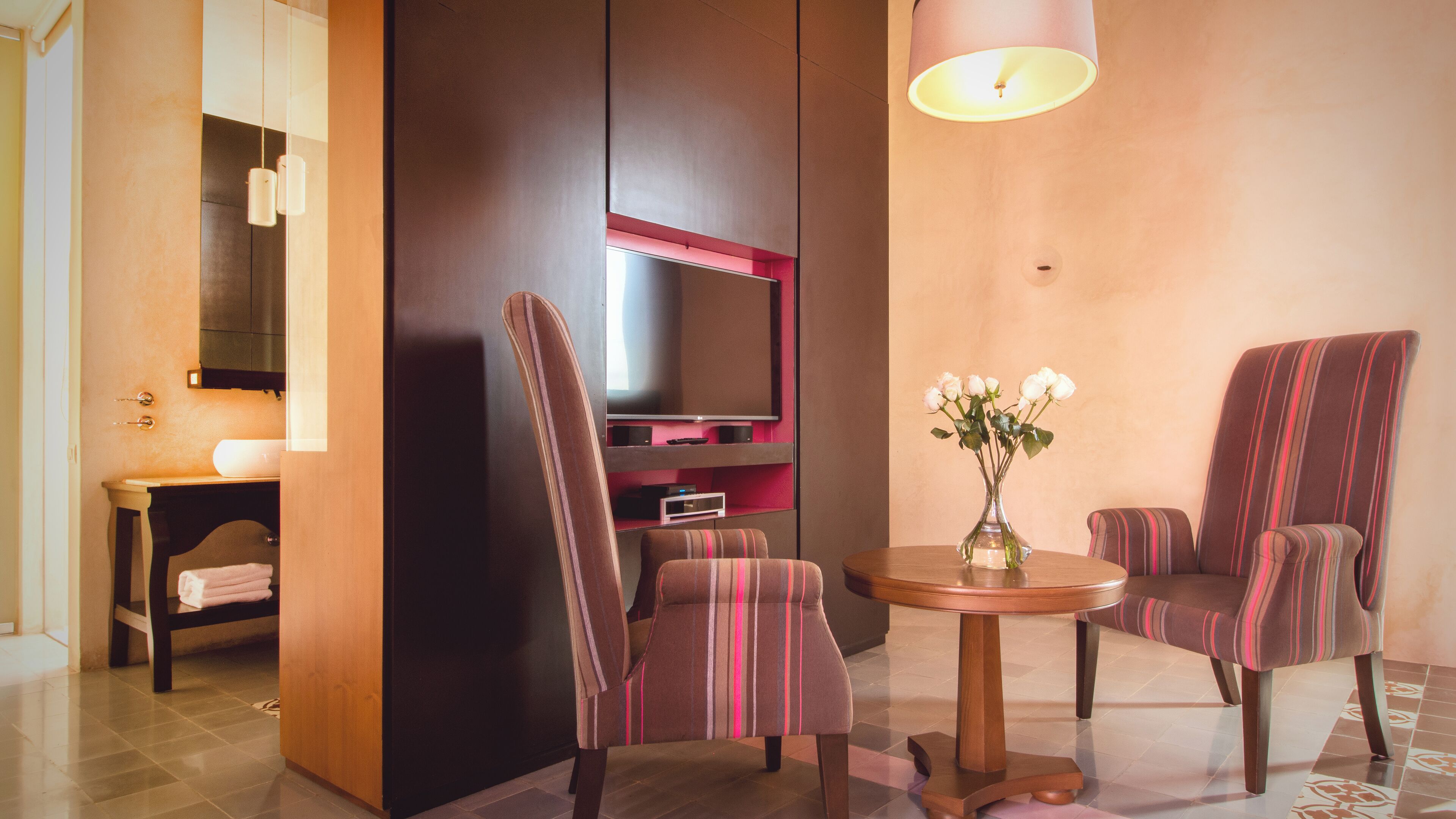 A modern hotel room with a flat-screen TV mounted on a dark wall, two striped armchairs, a small wooden table with a vase of flowers, and a lamp on the ceiling.