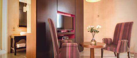 Una habitación de hotel moderna con un televisor de pantalla plana montado en una pared oscura, dos sillones a rayas, una pequeña mesa de madera con un jarrón de flores y una lámpara en el techo.