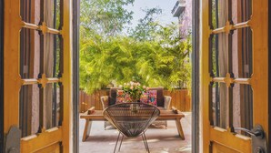 Una puerta de madera abierta que da a un patio con una mesa de madera, una silla de mimbre y un sofá con cojines de colores.