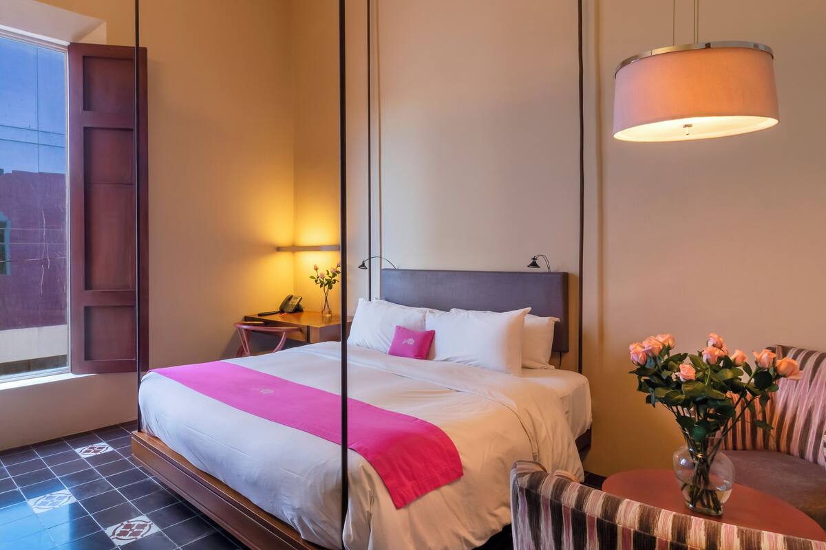 A hotel room with a large bed, a desk, a chair, a bedside table with a lamp, a vase with flowers, and a window with a view of buildings.