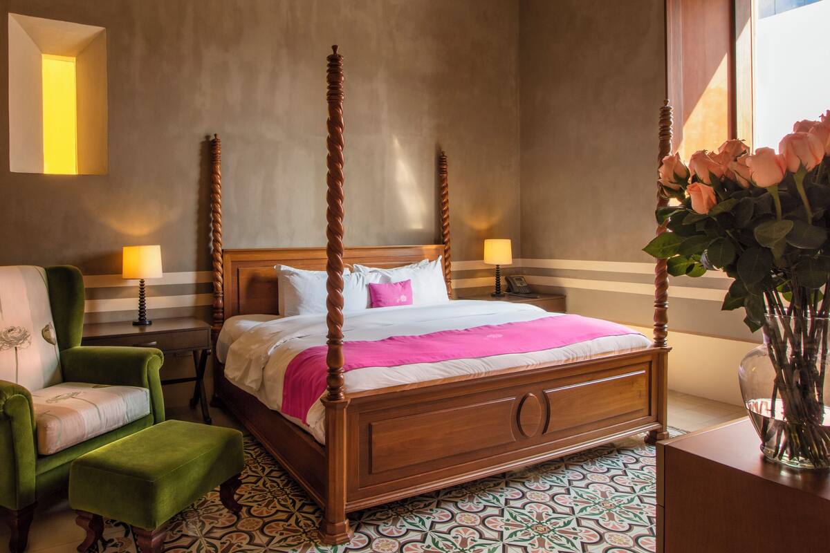 A four-poster bed with a wooden frame, a green armchair, a bedside table with a lamp, and a vase with flowers.