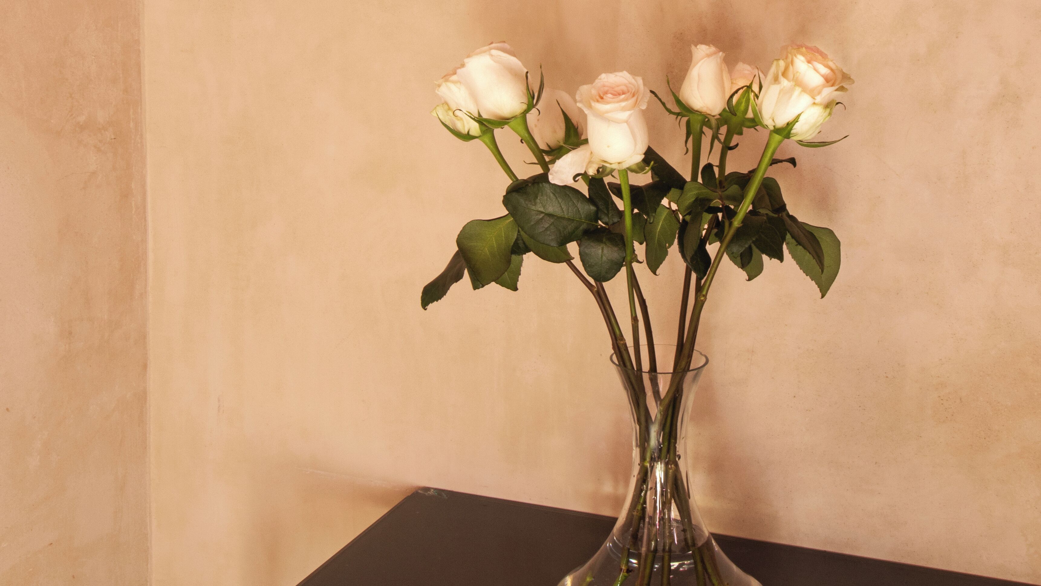 Un florero de cristal transparente con rosas blancas y hojas verdes sobre una superficie negra frente a una pared color crema.