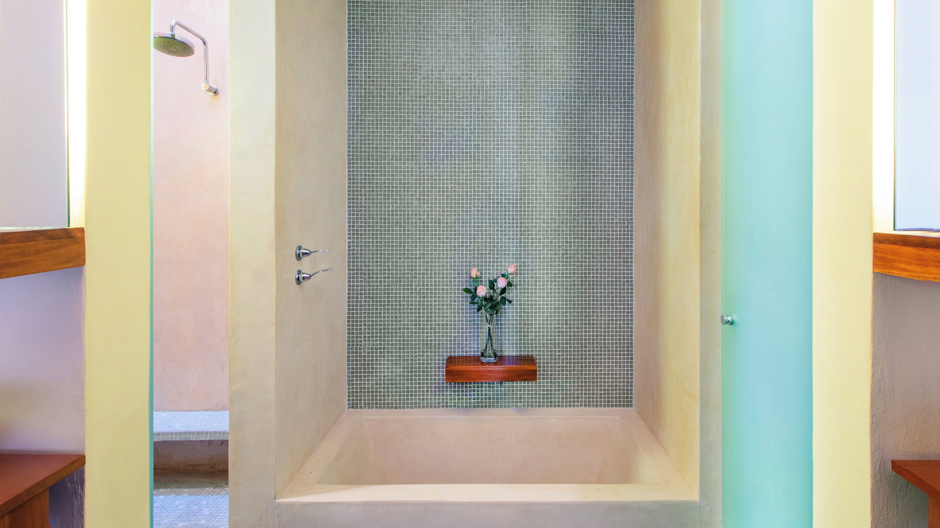 A bathroom with a tiled shower wall, a wooden shelf with a vase of flowers, and a door with a light switch.