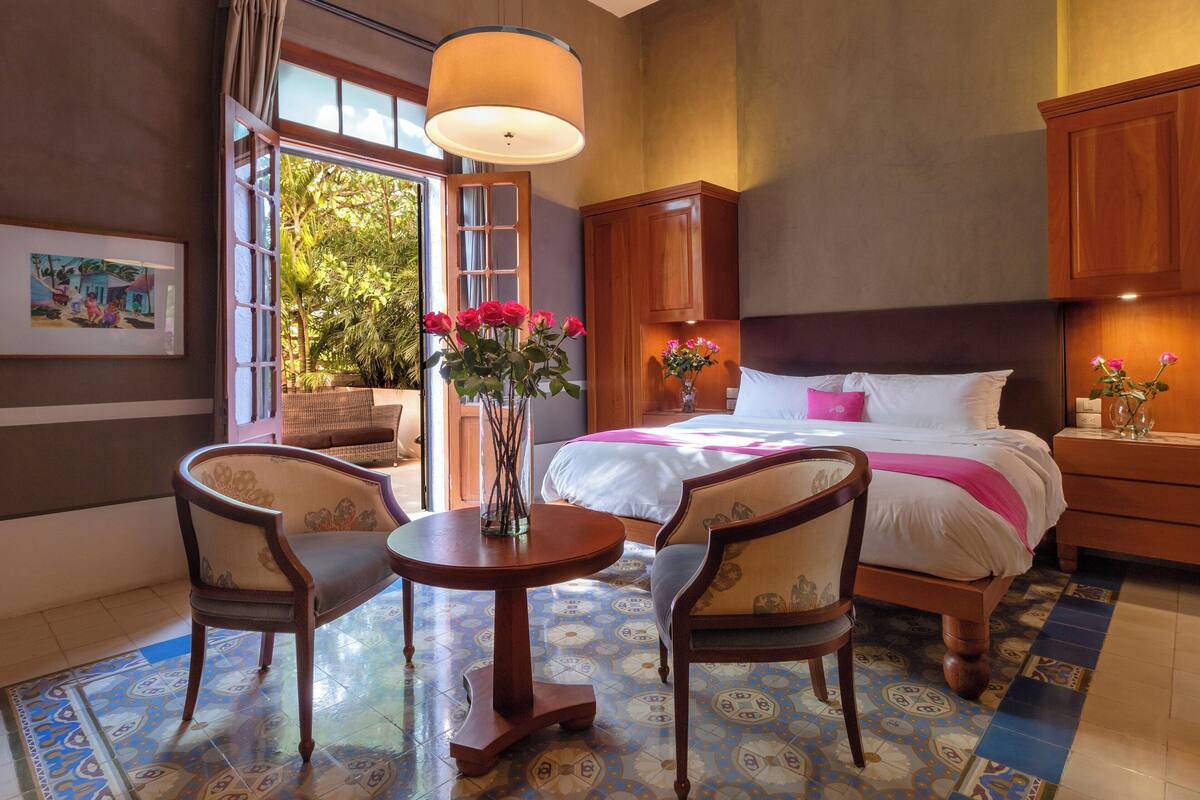 A hotel room with a bed, two chairs, a table, a vase with flowers, and a window with a view of greenery.