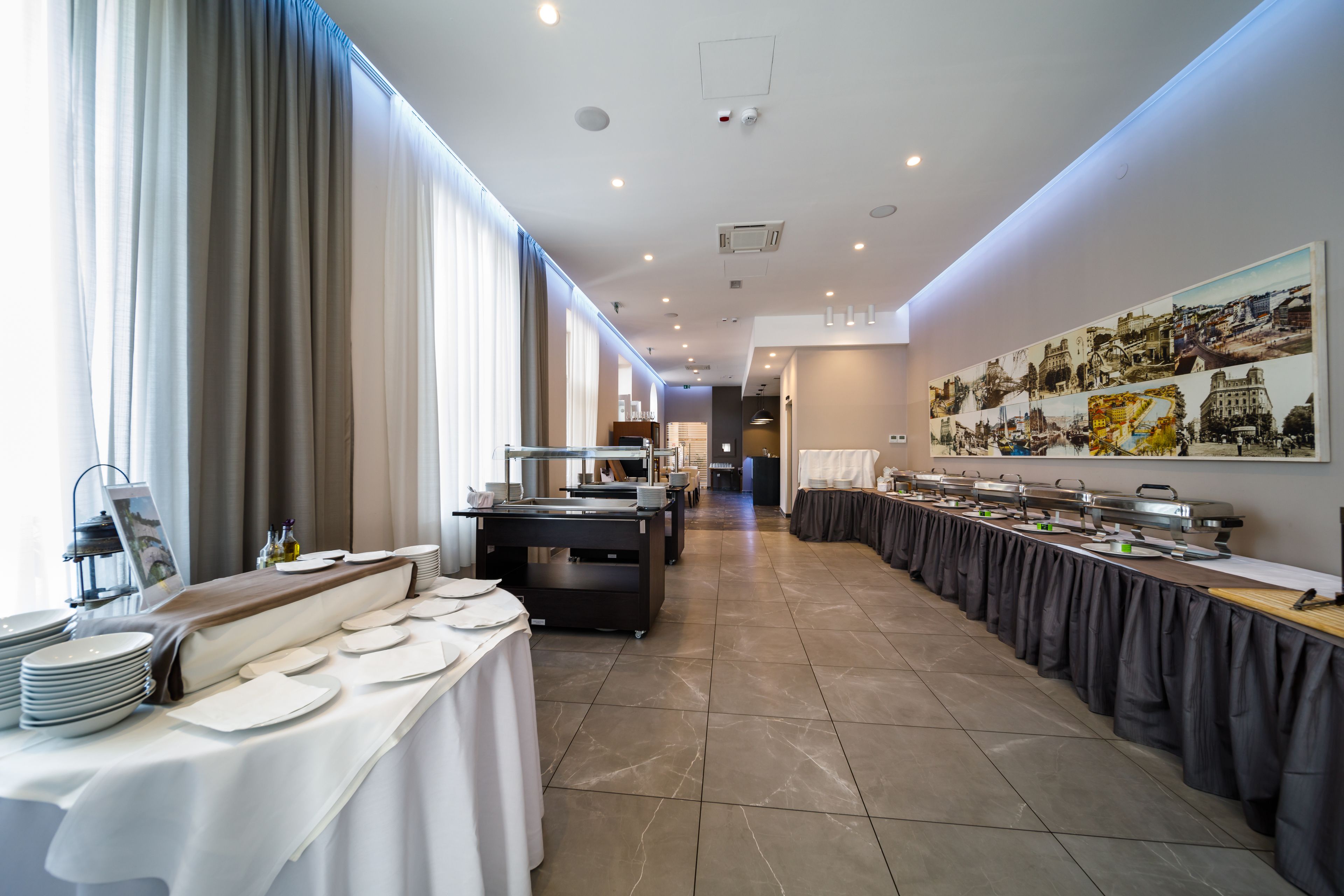 A modern hotel dining area with long tables, buffet stations, and a large cityscape mural on the wall.