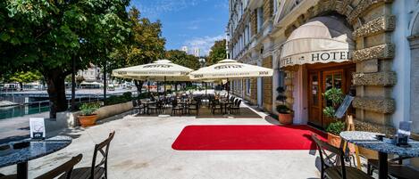 An outdoor seating area with tables and chairs, umbrellas, and a red carpet leading to a hotel entrance.