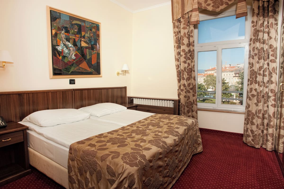 Habitación de hotel con una cama grande, una mesita de noche, un cuadro en la pared, una ventana con cortinas y vistas a edificios.