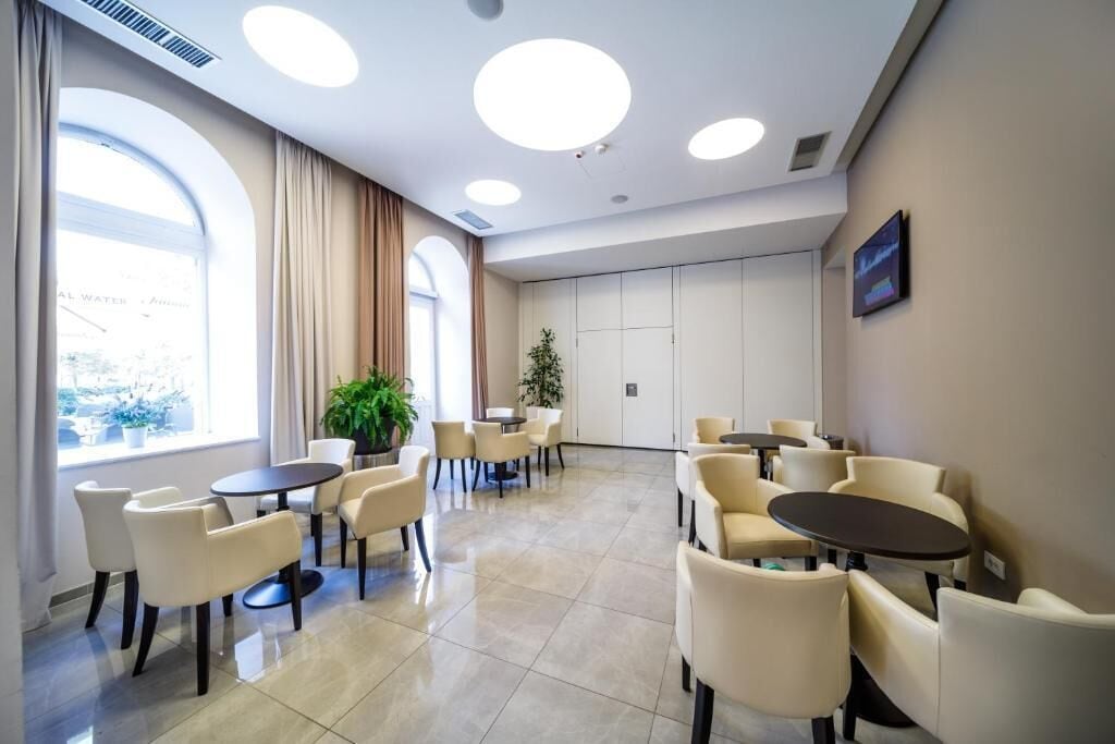 Et hotellobby med runde borde og stole, store vinduer med gardiner og en potteplante.
