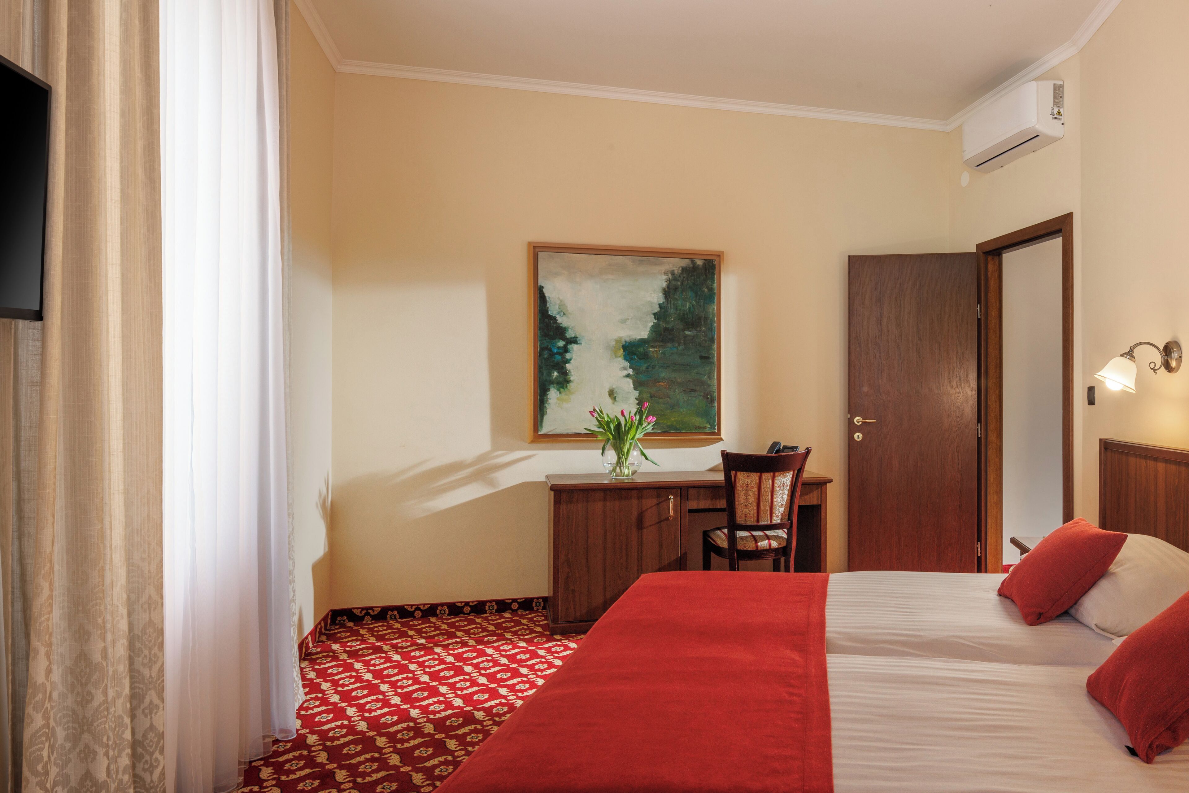 A hotel room with a bed, a desk, a chair, a painting, and an air conditioning unit.