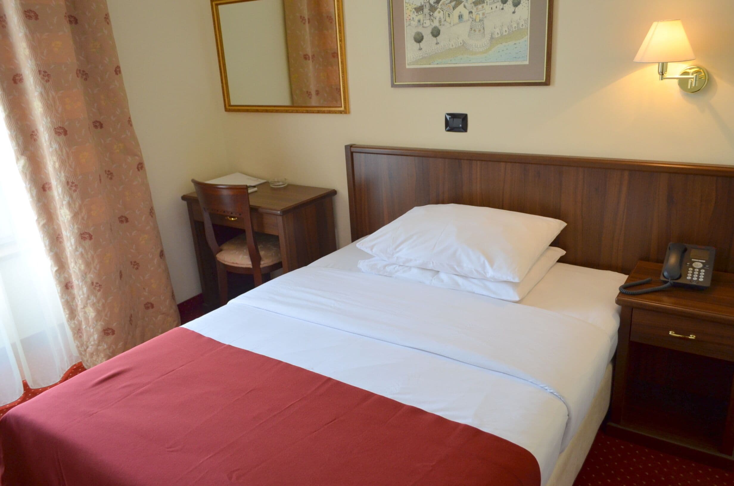 A hotel room with a bed, a bedside table with a lamp, a telephone, a chair, and a desk.