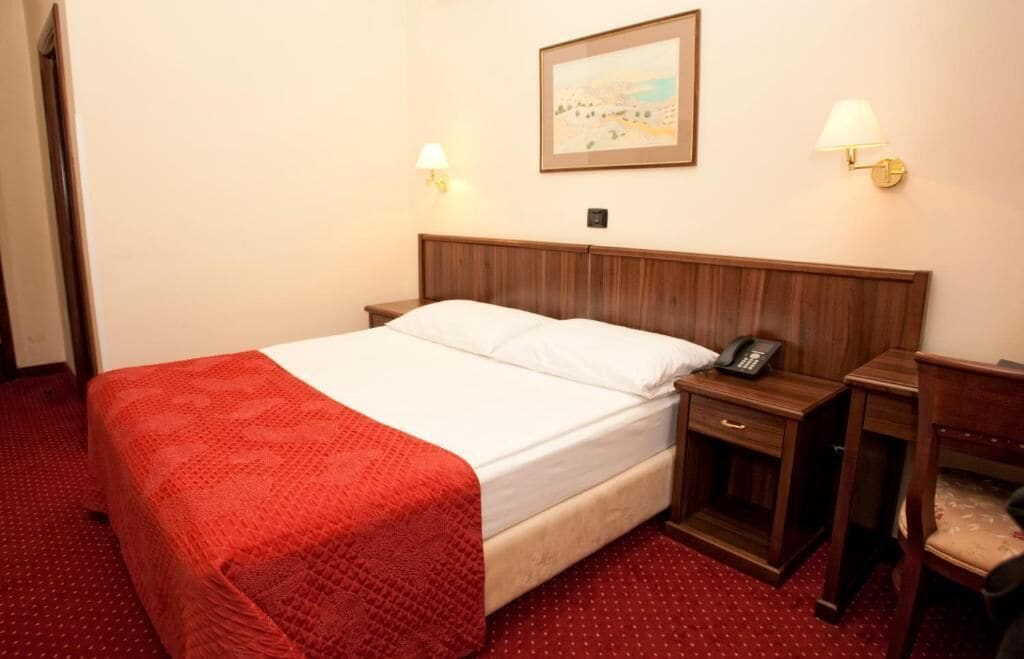 A hotel room with a wooden bed, a red blanket, a bedside table with a lamp, and a painting on the wall.