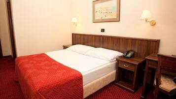 A hotel room with a wooden bed, a red blanket, a bedside table with a lamp, and a painting on the wall.