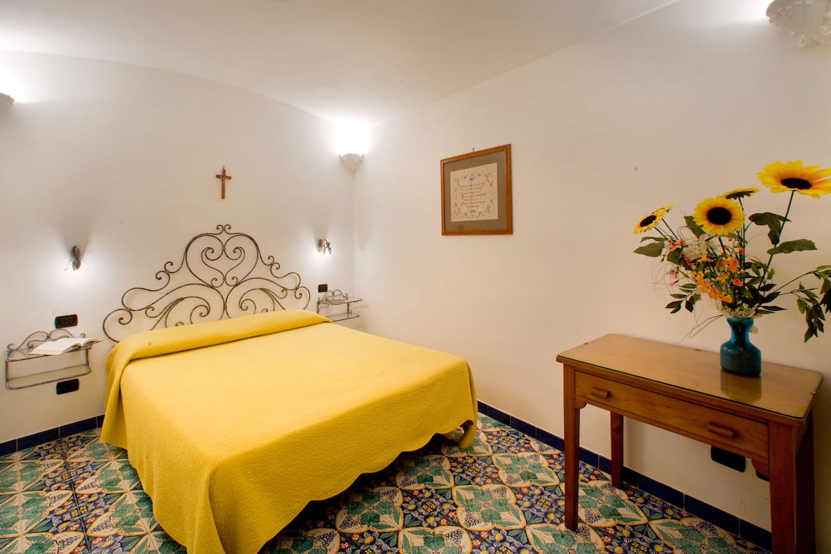 A bedroom with a bed, a nightstand, a bedside table, a wall-mounted cross, a framed certificate, and a vase with flowers.