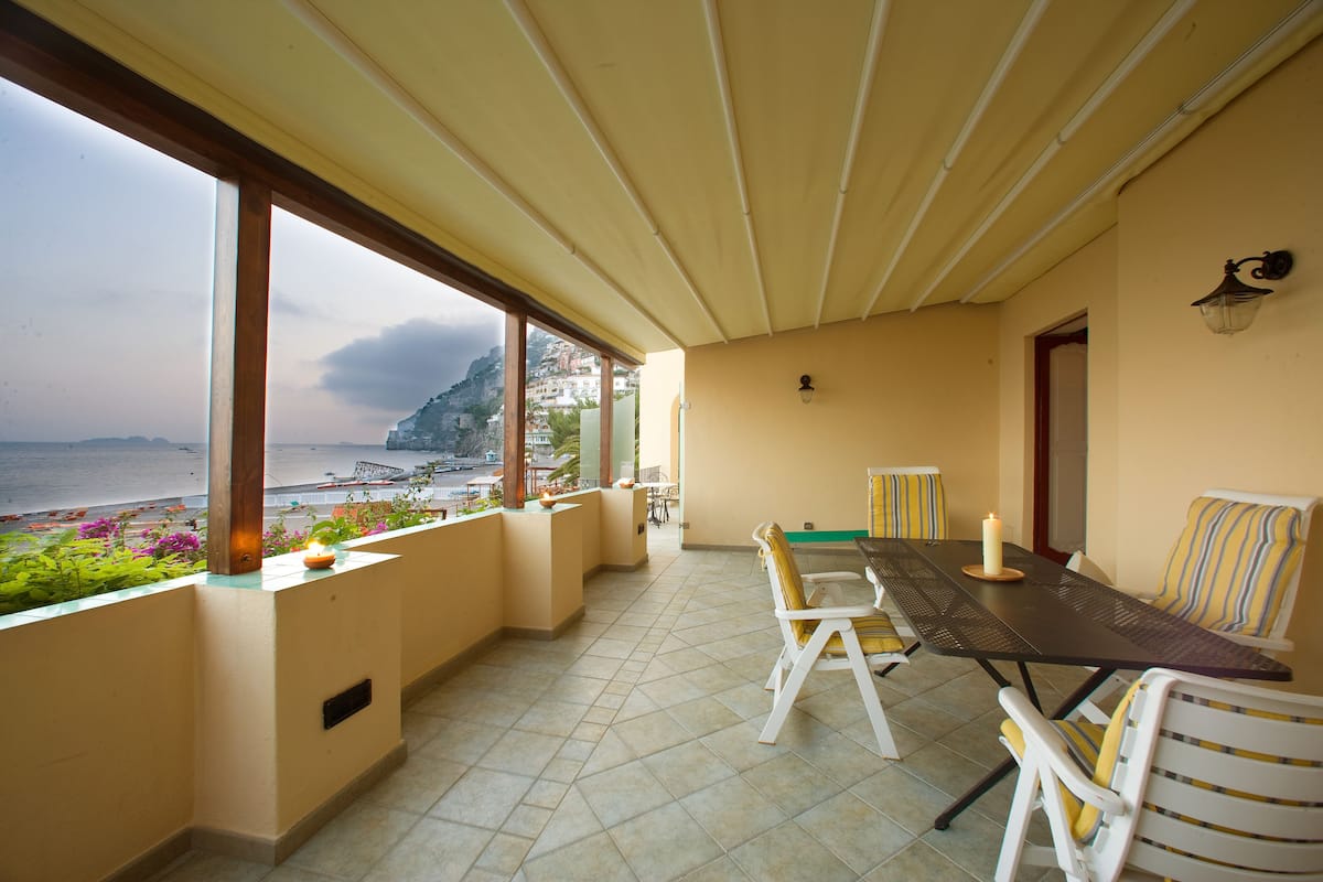 A covered outdoor dining area with a table and chairs, a view of the sea, and a balcony with plants.