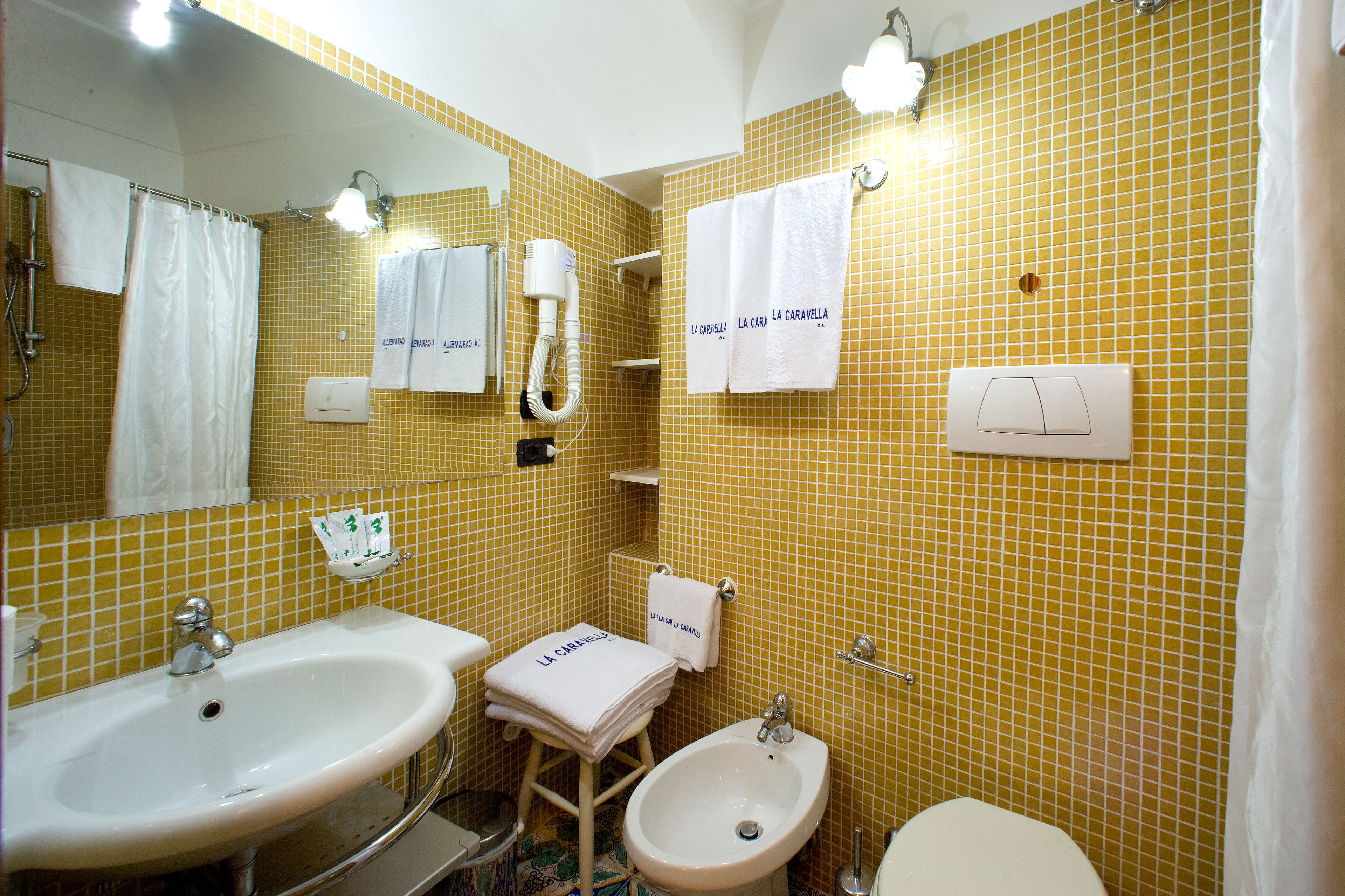 Un baño con paredes de azulejos amarillos, dos lavamanos, un bidé y varias toallas blancas con texto.