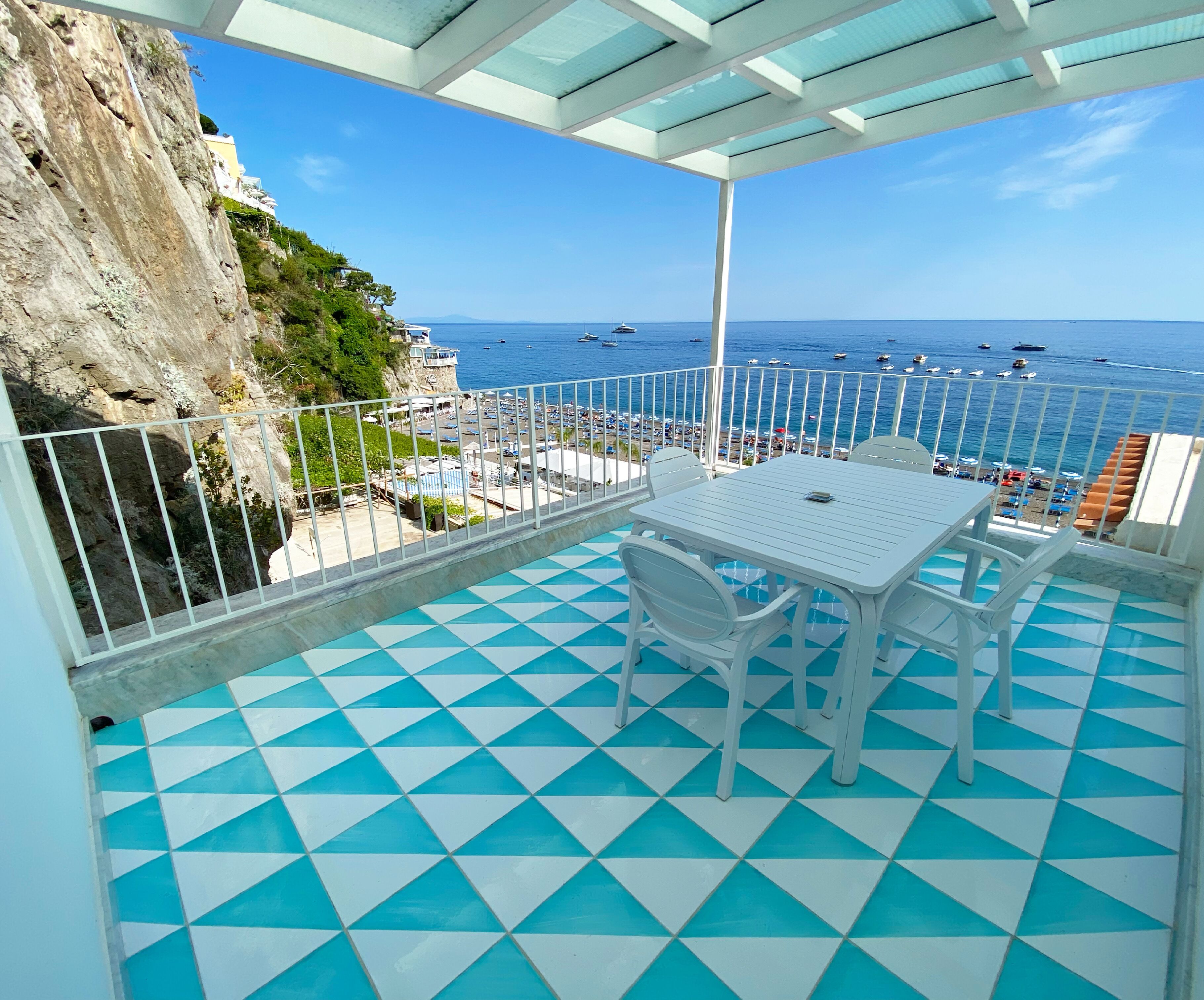 A balcony with a checkered floor, white furniture, and a view of the sea and beach.
